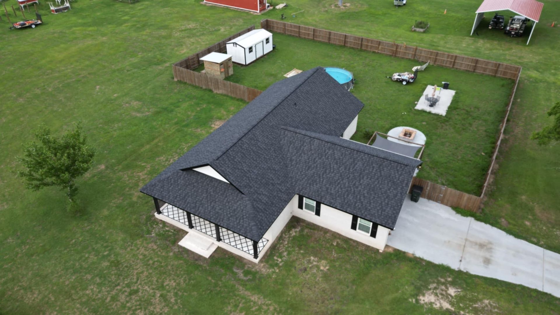 Aerial view of a house with black roof, fenced backyard, and a long concrete driveway.