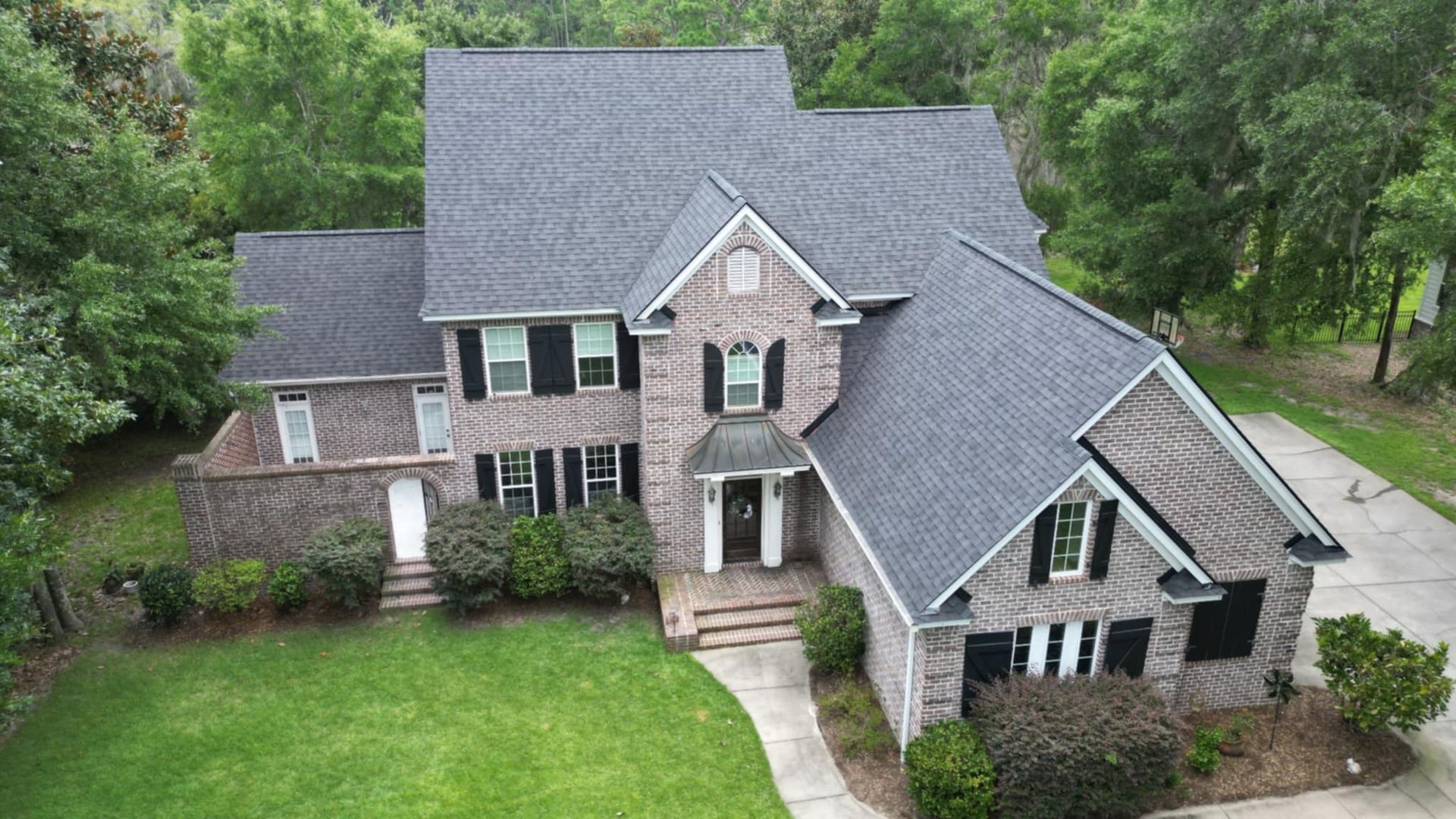 Large brick house with gray roof and shutters, surrounded by green lawn and trees.
