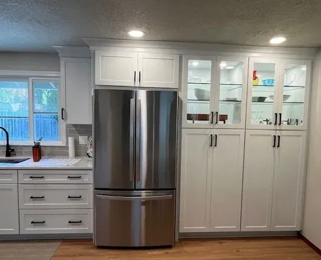 A stainless steel refrigerator is flanked by white kitchen cabinets, some with glass doors and interior lighting.