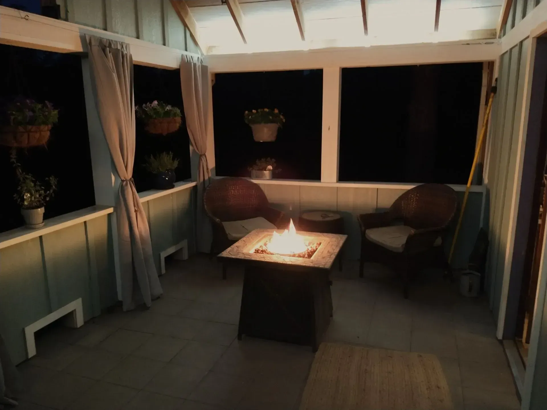 A lit fire pit in an enclosed porch with chairs and hanging plants, viewed at night.