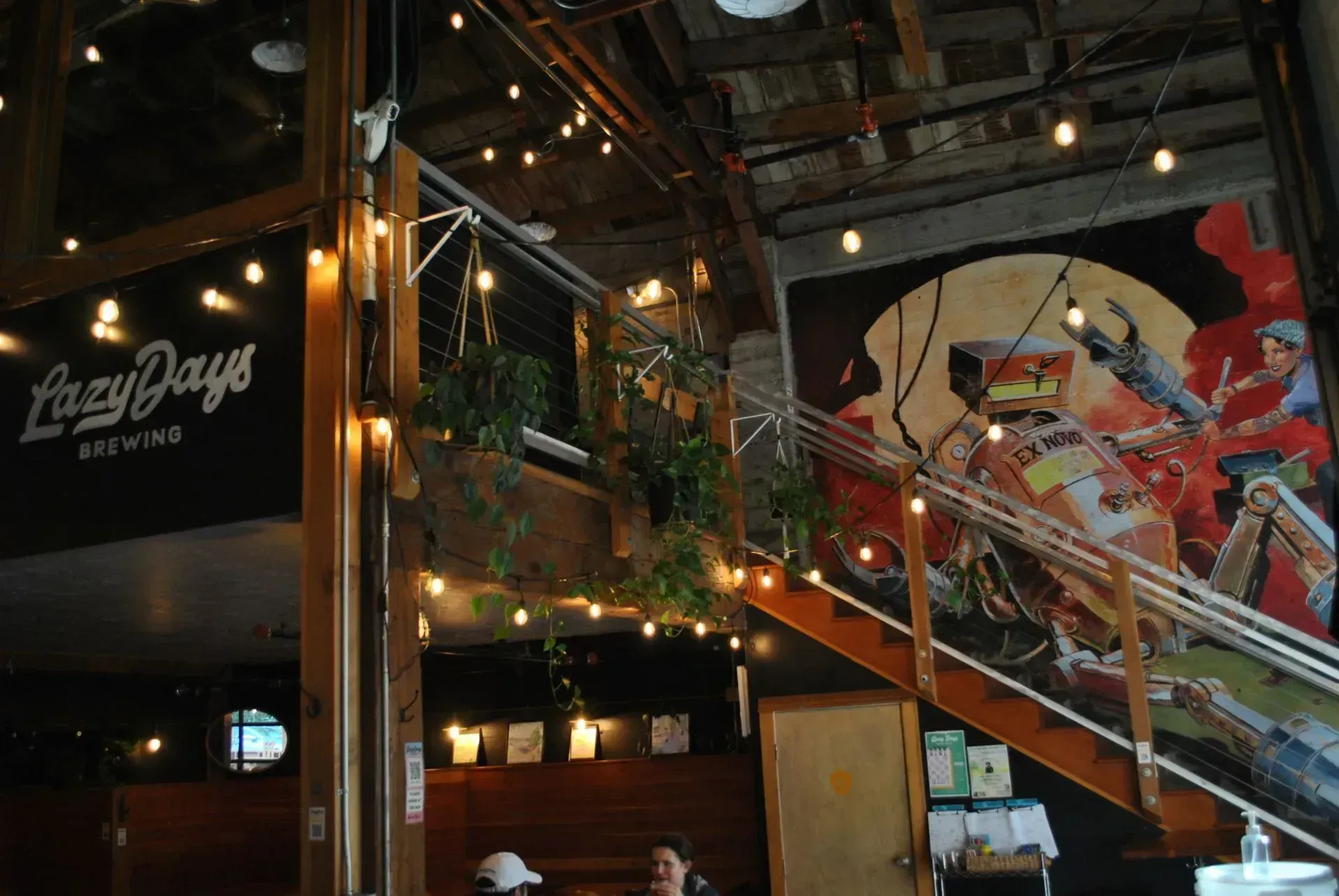Interior of Lazy Days Brewing with string lights, mural, staircase, and hanging plants.