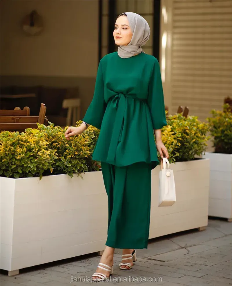 A woman wearing a hijab and a green dress is standing in front of a potted plant.