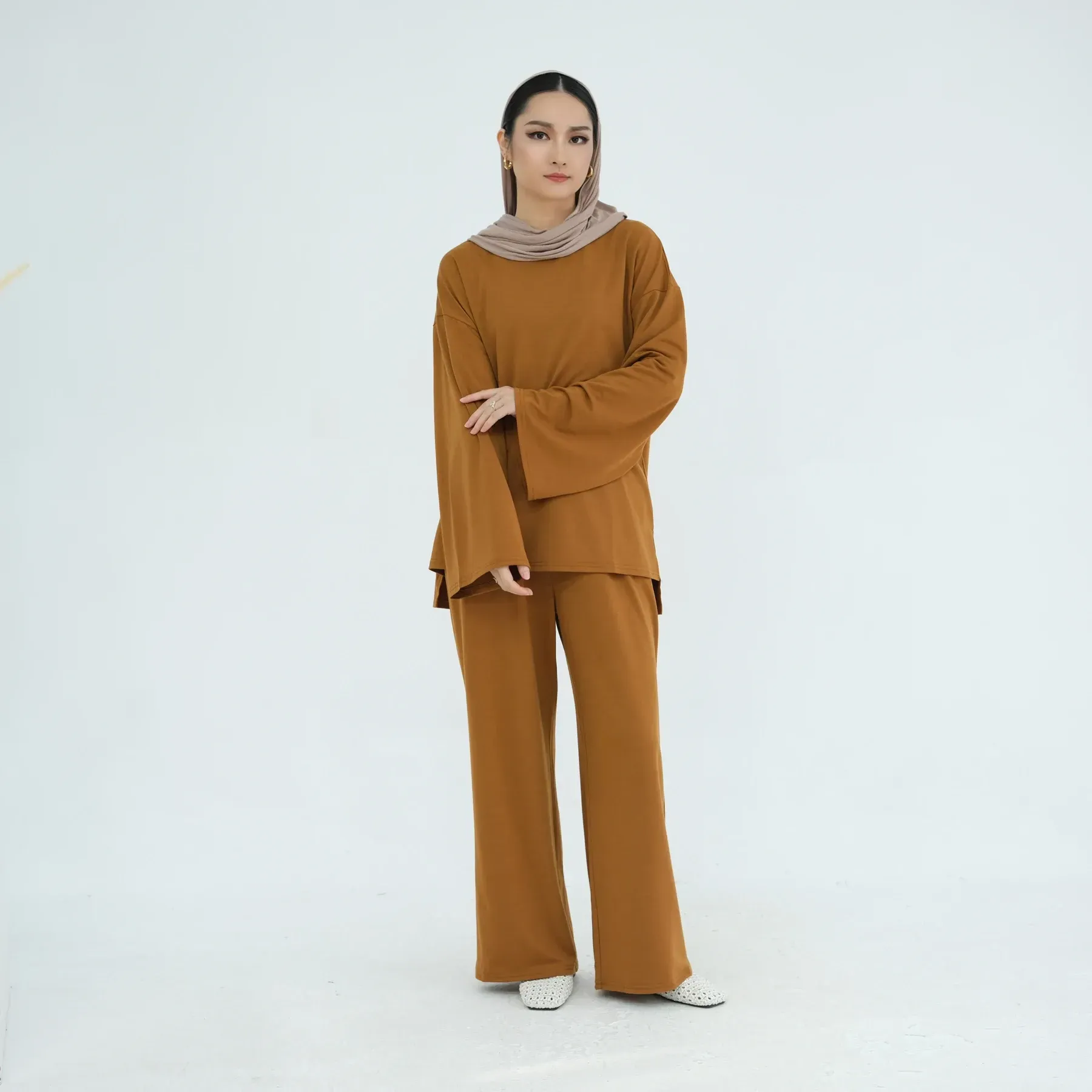 A woman wearing a hijab and a brown shirt and pants is standing on a white background.