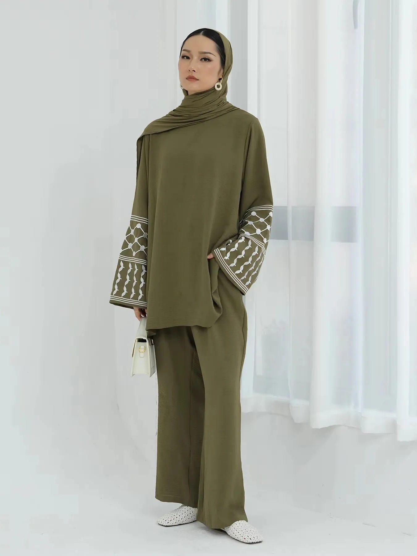 A woman wearing a hijab and a green dress is standing in front of a window.