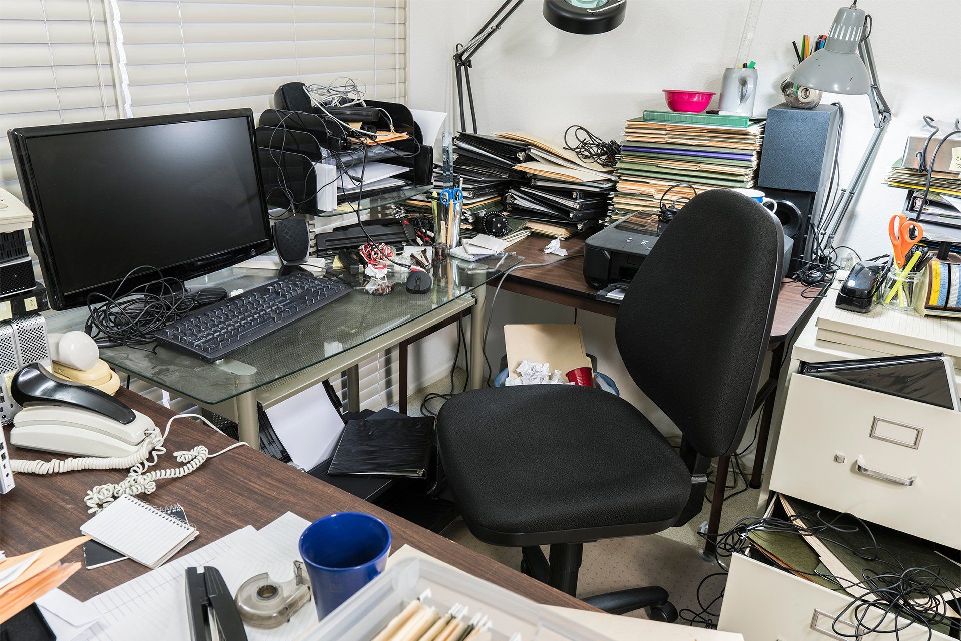 A messy office with a desk , chair , computer , printer , and other items.