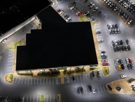 Aerial view of a large dark-roofed building surrounded by a parking lot with many cars at night.