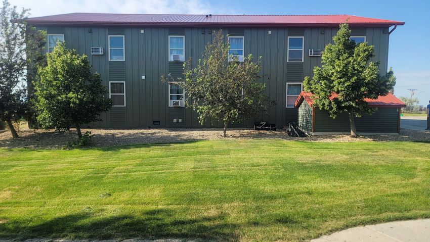 Two-story building with green siding and a red roof, fronted by a grassy lawn and small trees under a blue sky.