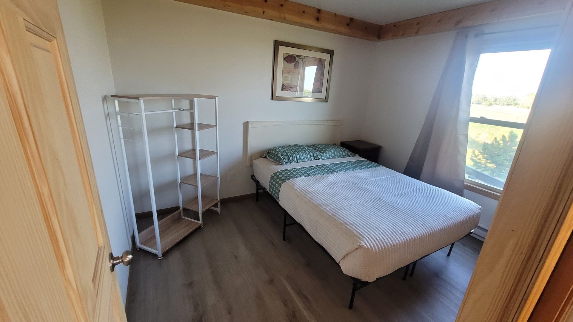 Bedroom with a bed, shelf unit, and window. Pale walls, wood trim, and a small painting are visible.
