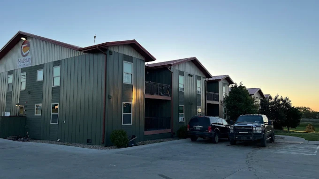 Two-story green metal-clad building with a parking lot. Several vehicles are parked in front.