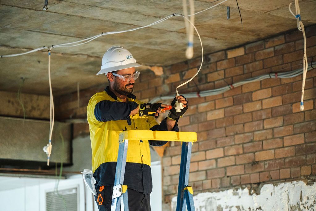 Electrician in safety gear works on wiring, standing on a step stool in a construction site with brick wall.