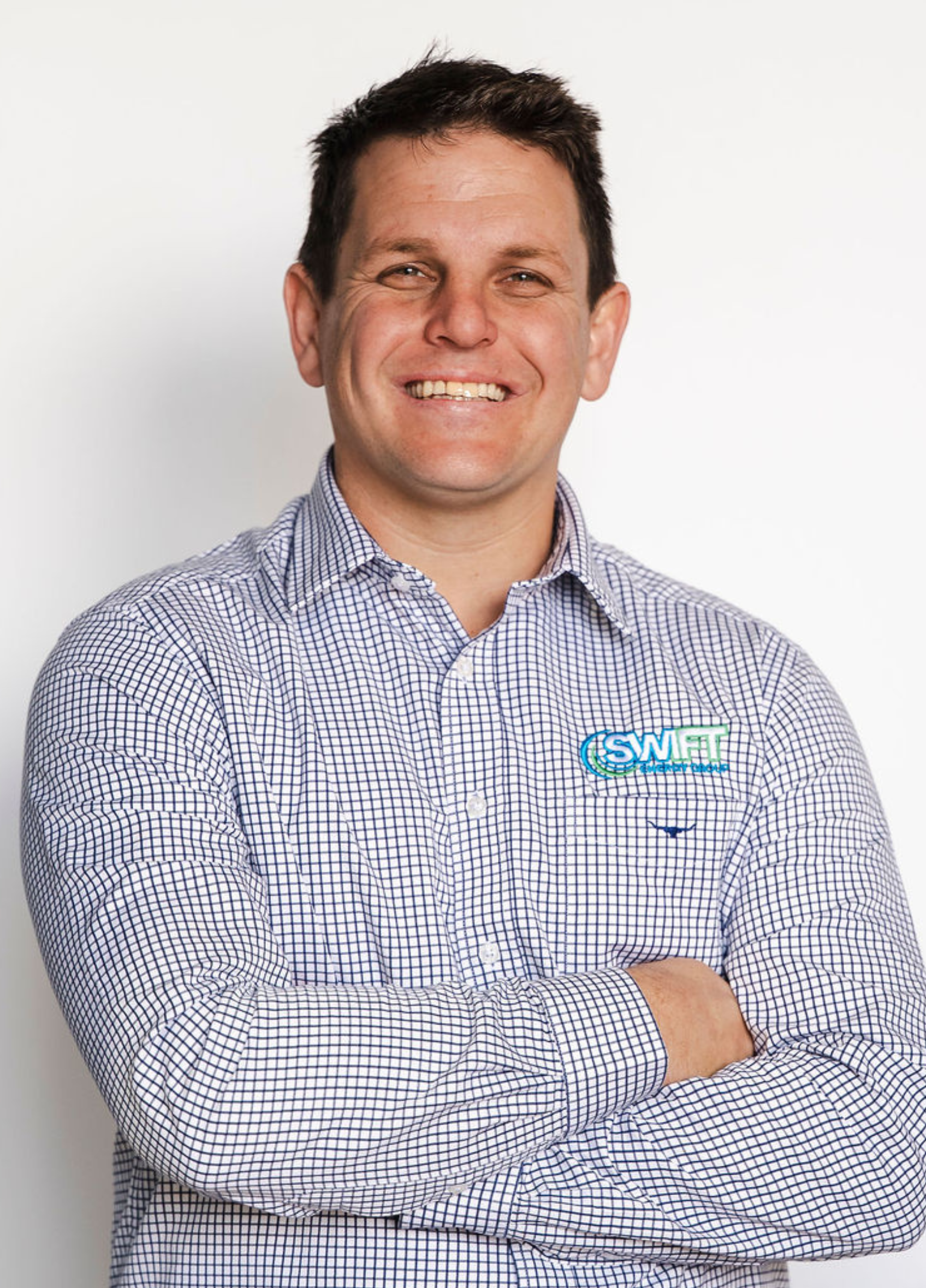 Man with arms crossed smiling, wearing blue checkered shirt, logo on left chest.