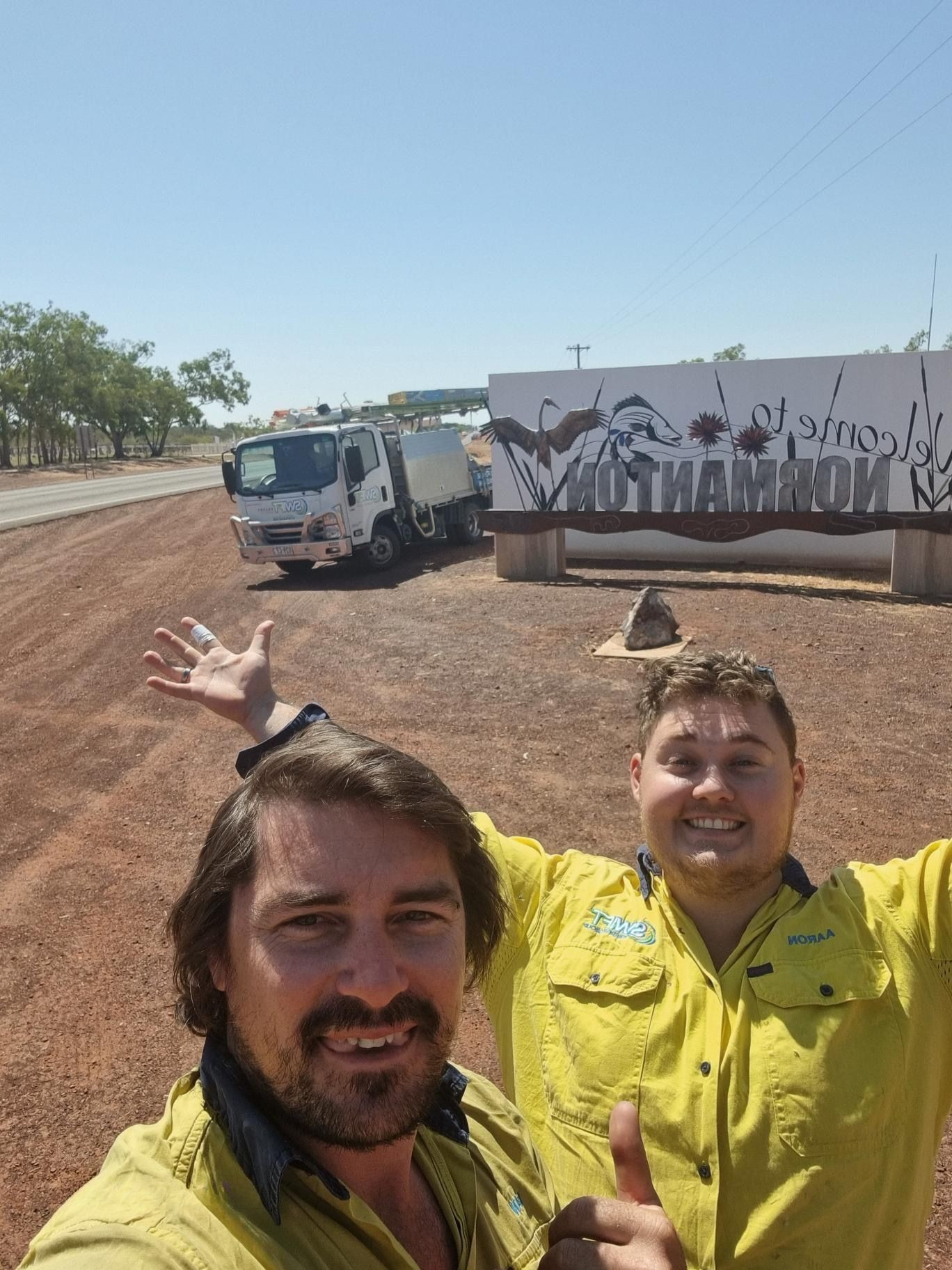 Two smiling people in yellow shirts pose beside a truck on a sunny dirt road.