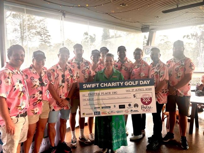 Group holding a “Swift Charity Golf Day” banner in a covered outdoor venue.