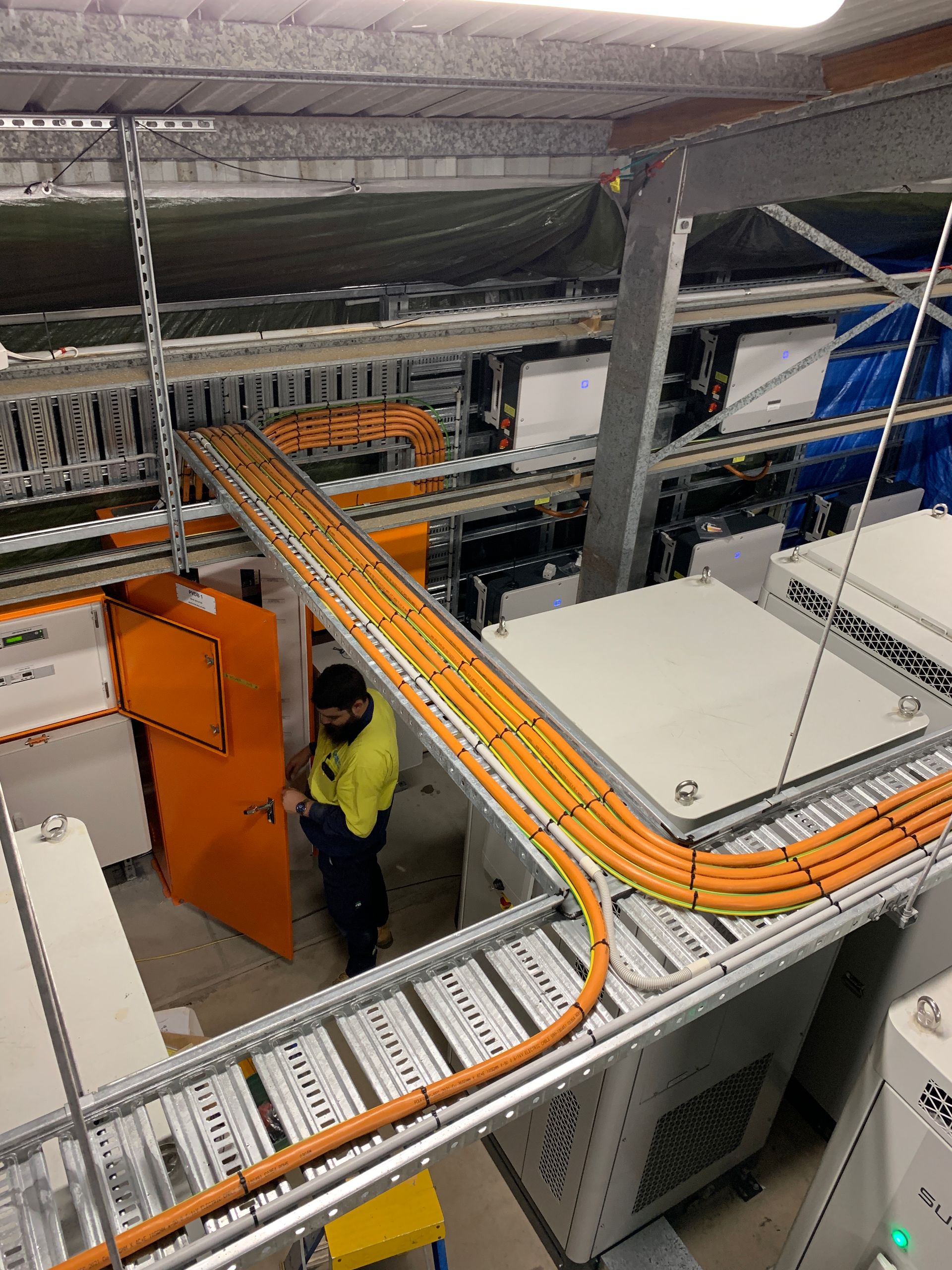 Person in a yellow shirt by an orange door, near orange cables on a metal cable tray in a technical setting.