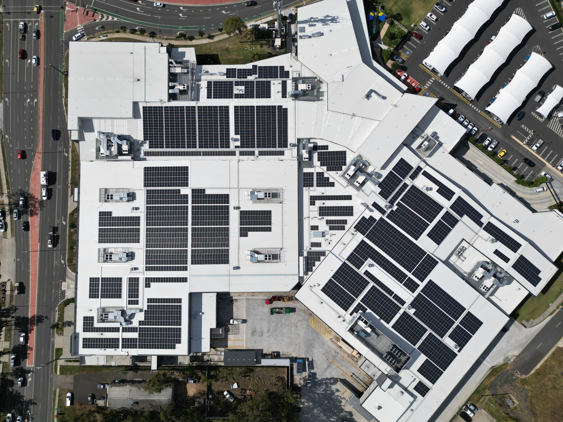 Overhead view of a large commercial building roof covered in solar panels, next to a parking lot and a road with cars.