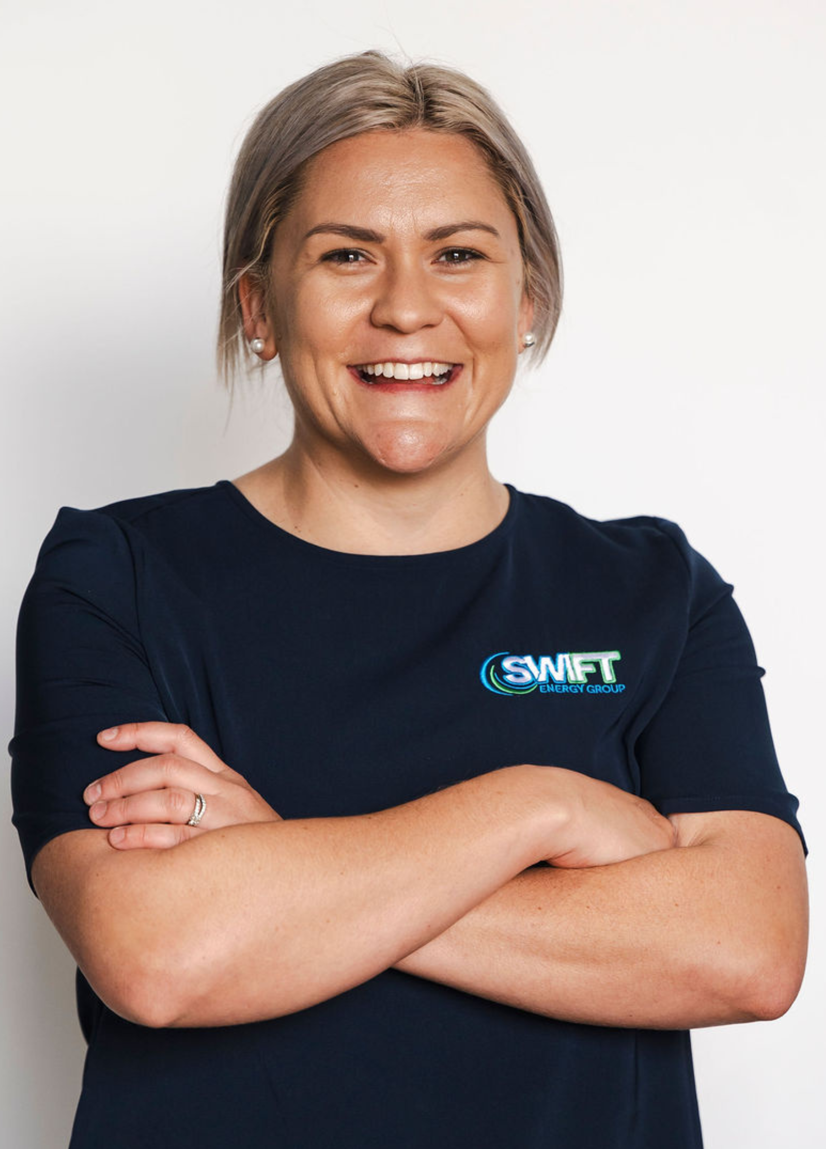 Woman with blonde hair, arms crossed, smiling, wearing a navy shirt with a logo, against a white backdrop.