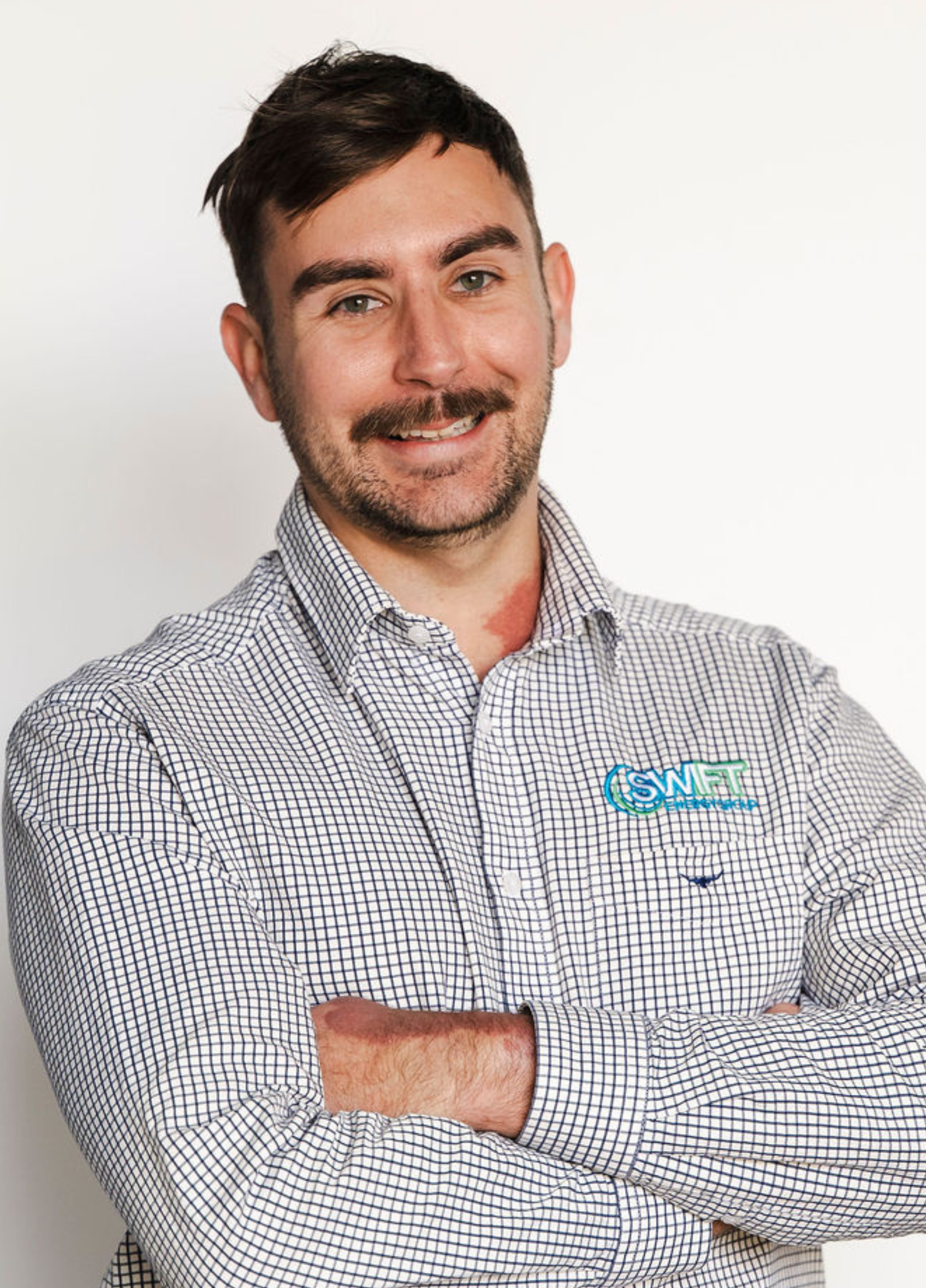 Man with mustache smiles, arms crossed, wearing a patterned button-up shirt in front of a white background.