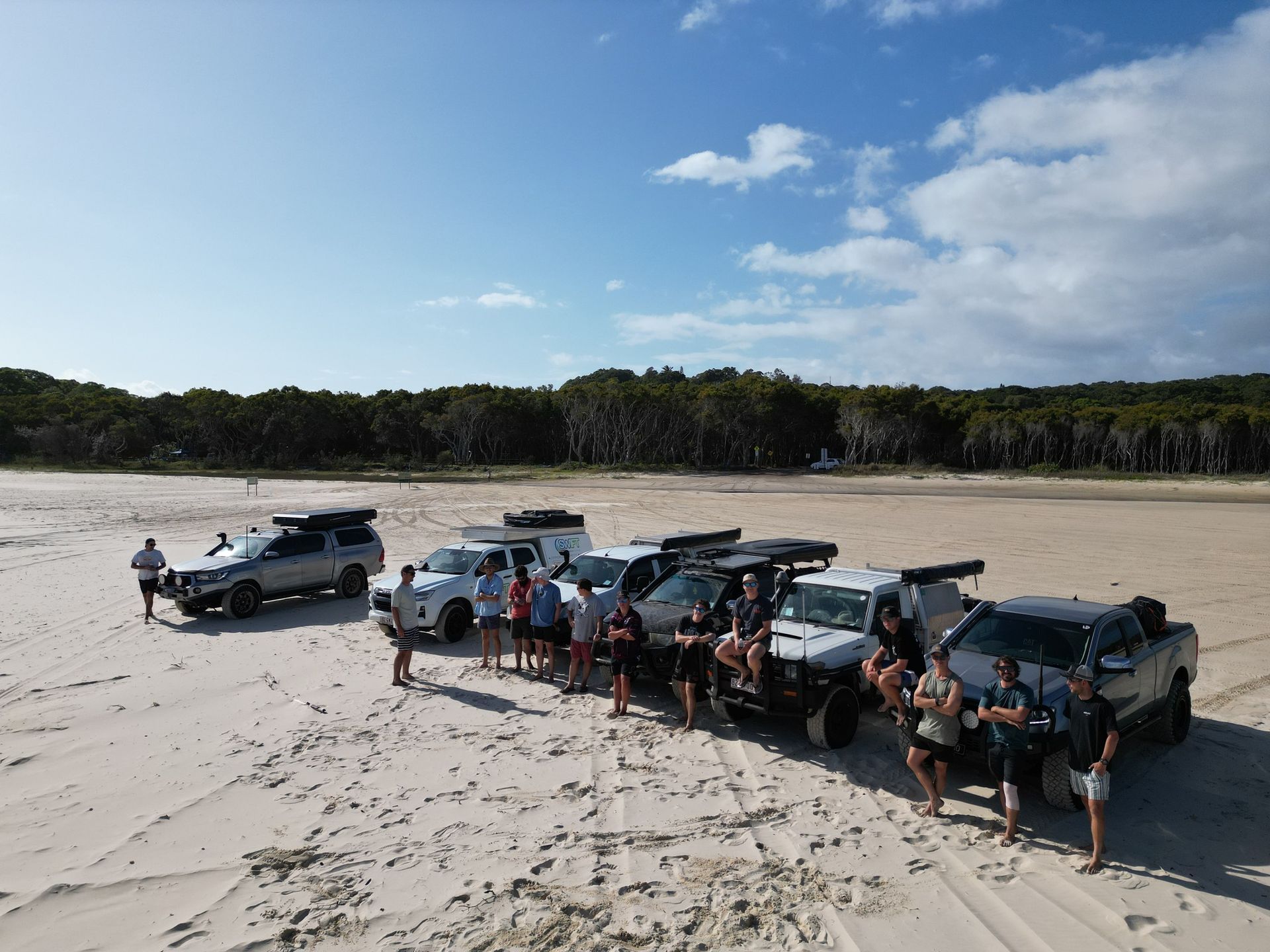A group of vehicles and people gathered on a sandy beach under a blue sky.