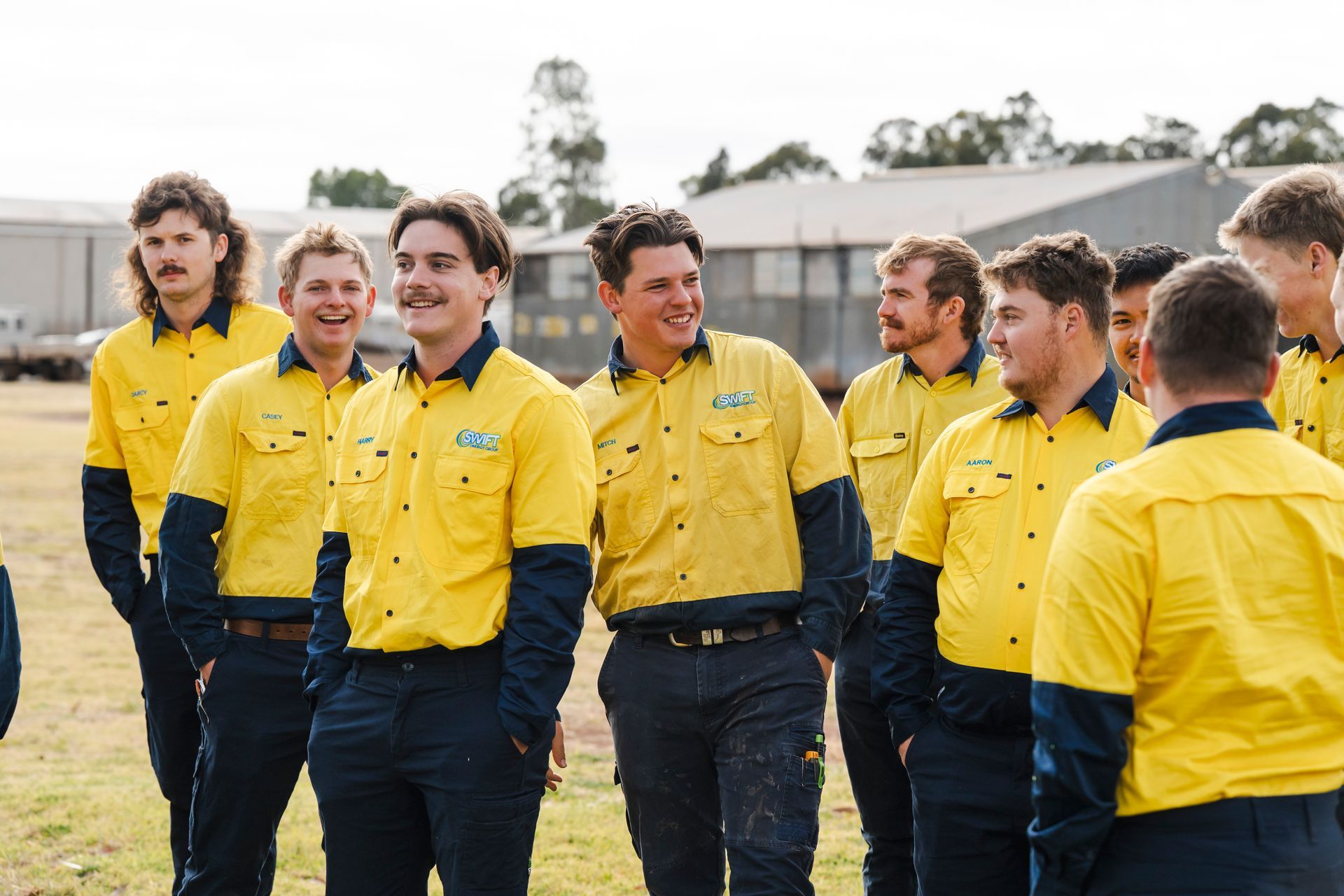 Three electricians in yellow shirts and blue pants smile outside.