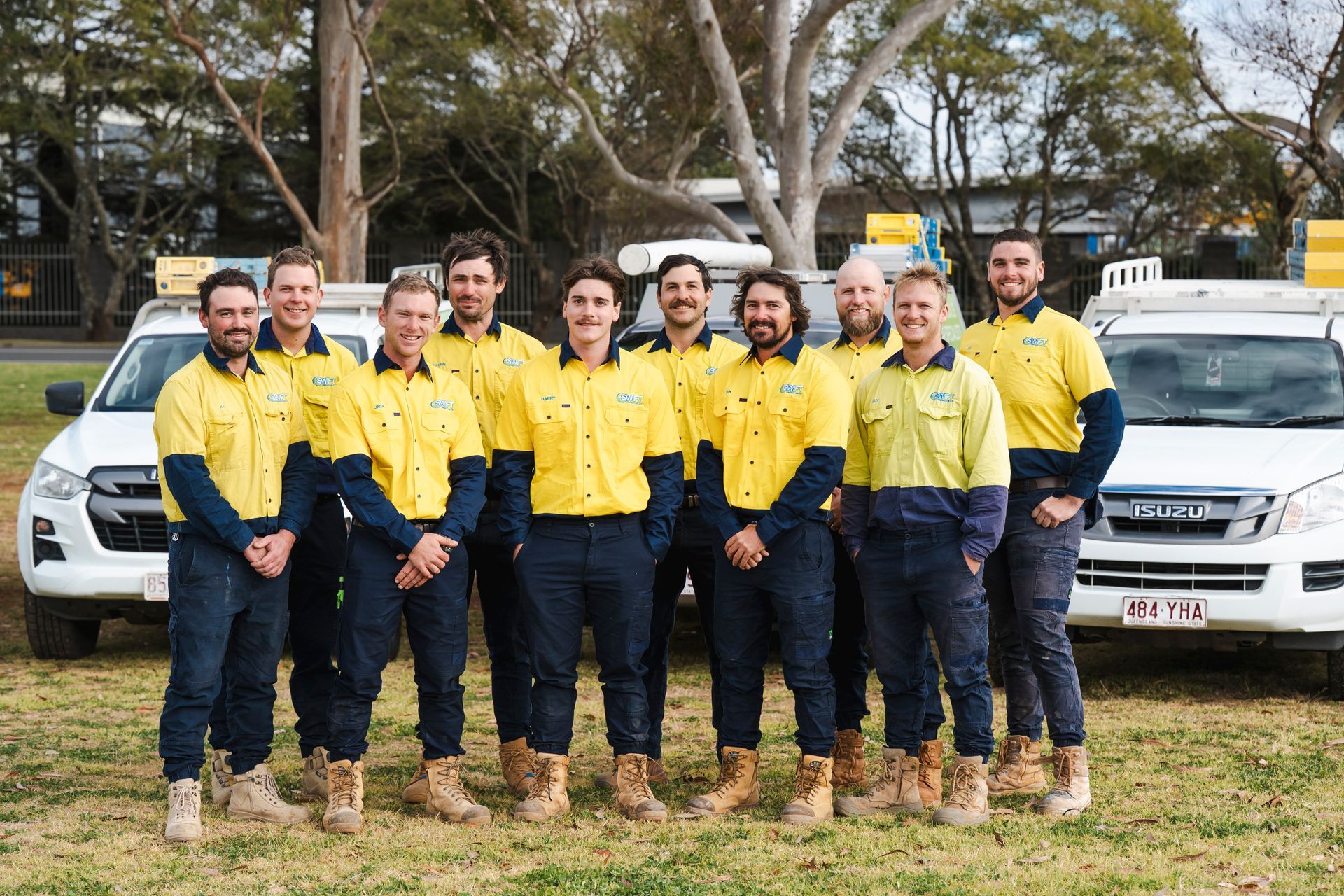 Group of workers in yellow shirts and blue pants, standing in front of two white work trucks.