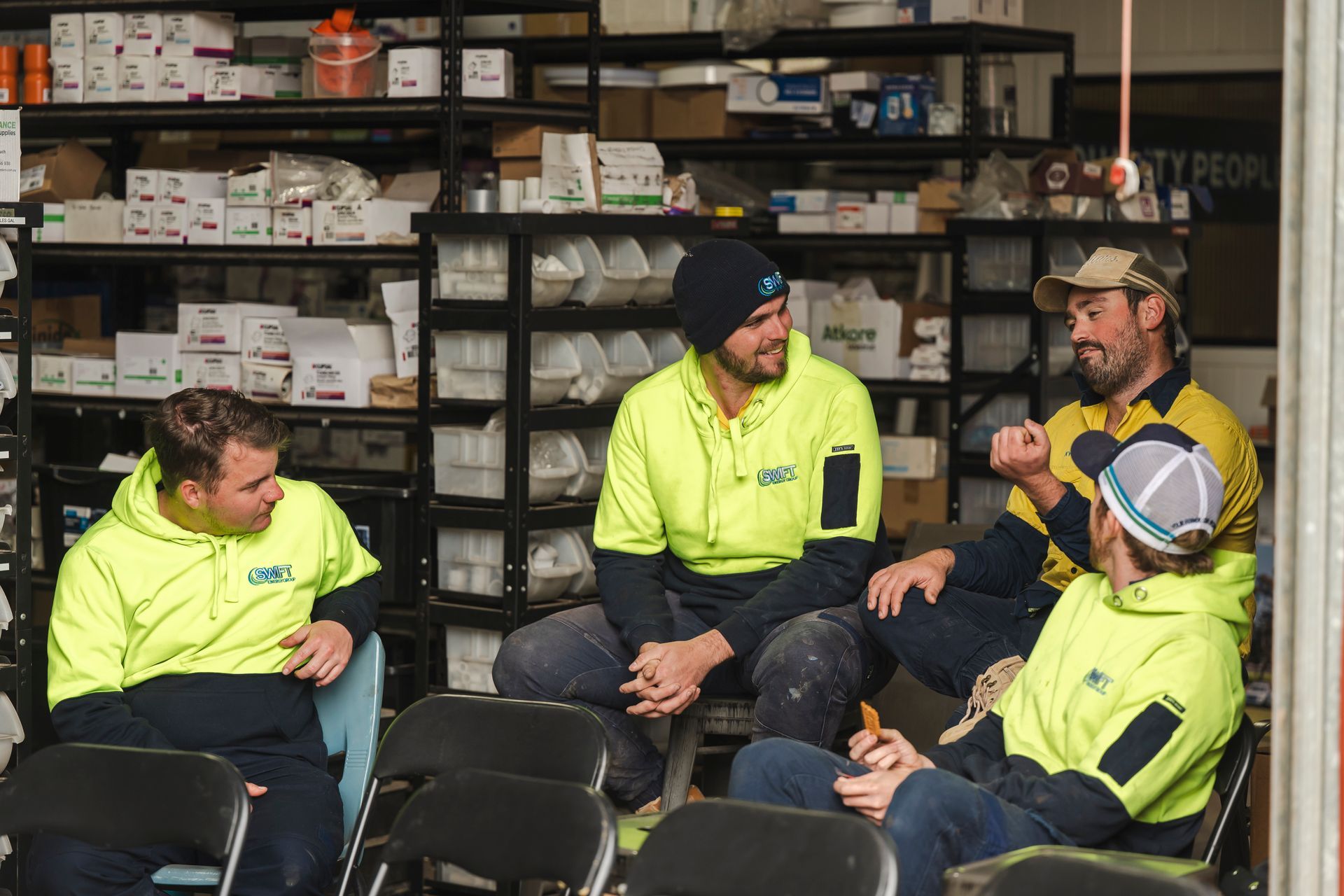 Workers in neon shirts and hard hats sit together in a warehouse, talking beside shelves of supplies.