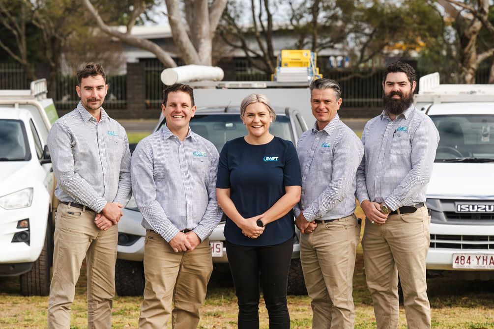 Five people posing in front of vehicles. Three men, one woman and one man. They wear work attire. Outdoors.