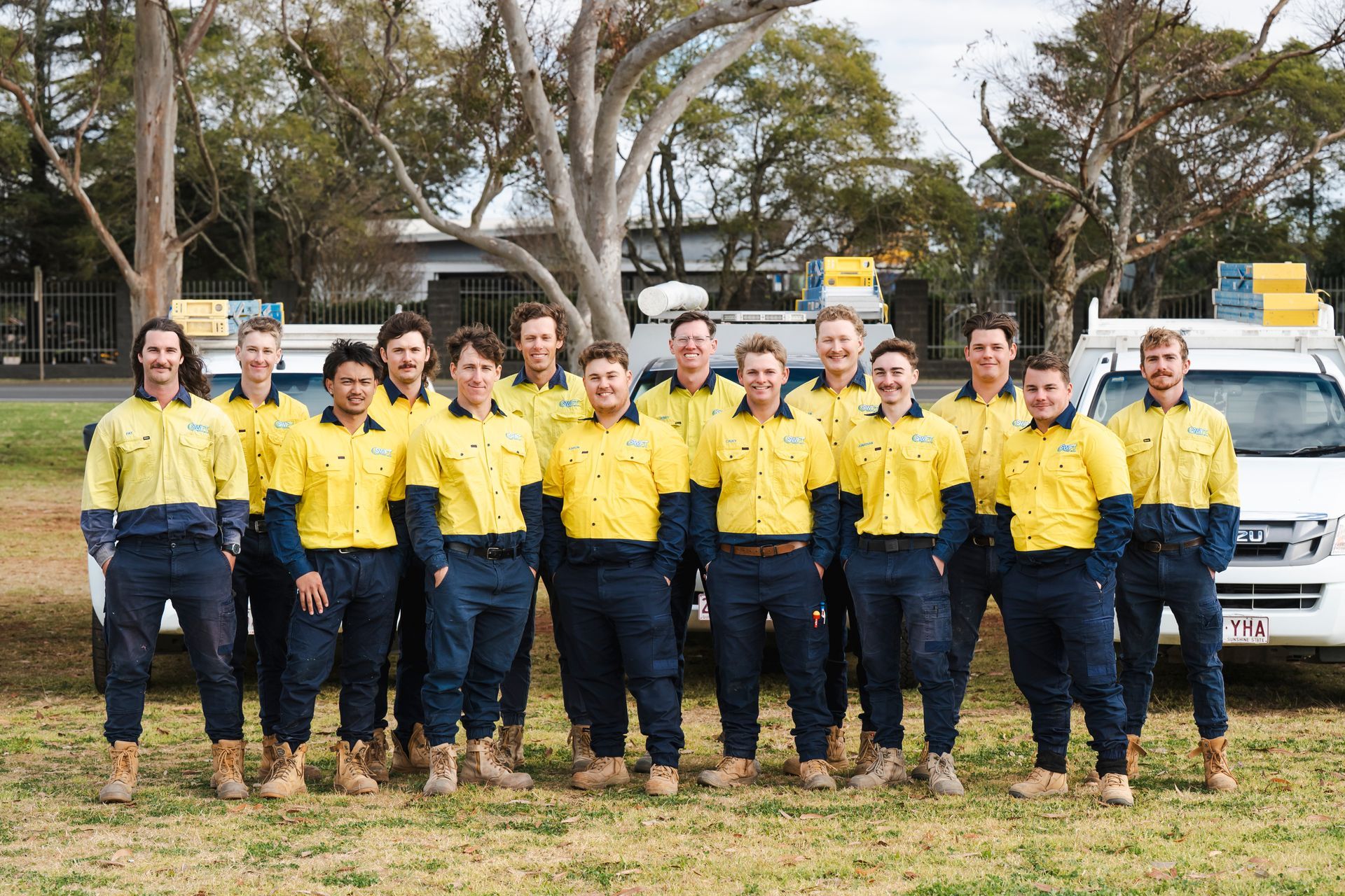 Group of workers in yellow and blue uniforms, standing outdoors in front of vehicles.