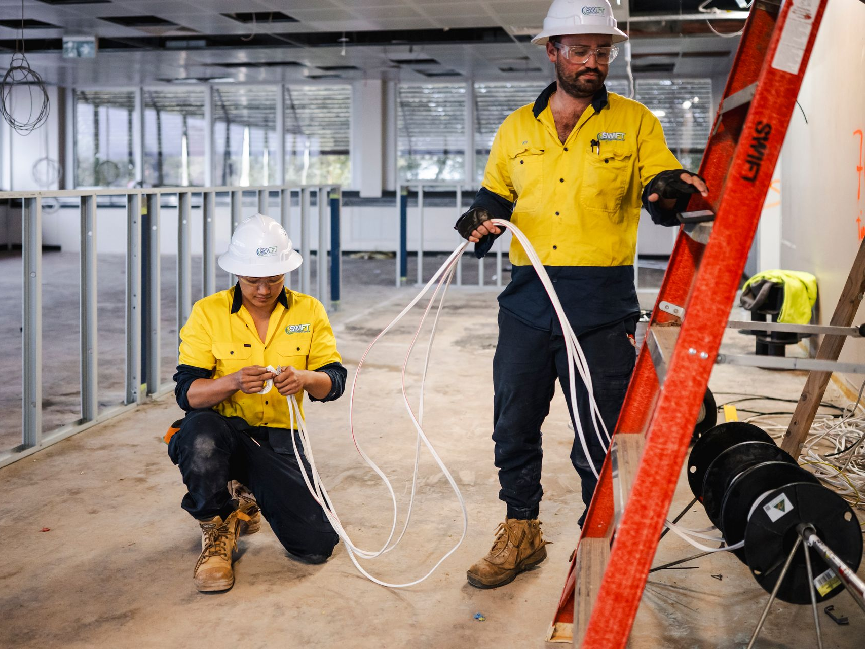 Two construction workers inside a building under construction, working with wires. One is on a ladder.