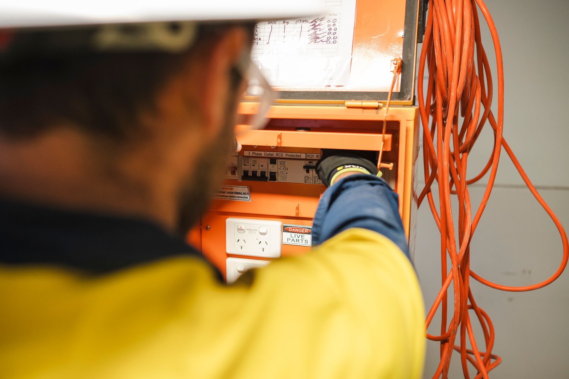 Electrician on a ladder connecting wires to ceiling. Wears hard hat, safety glasses, and work gloves.