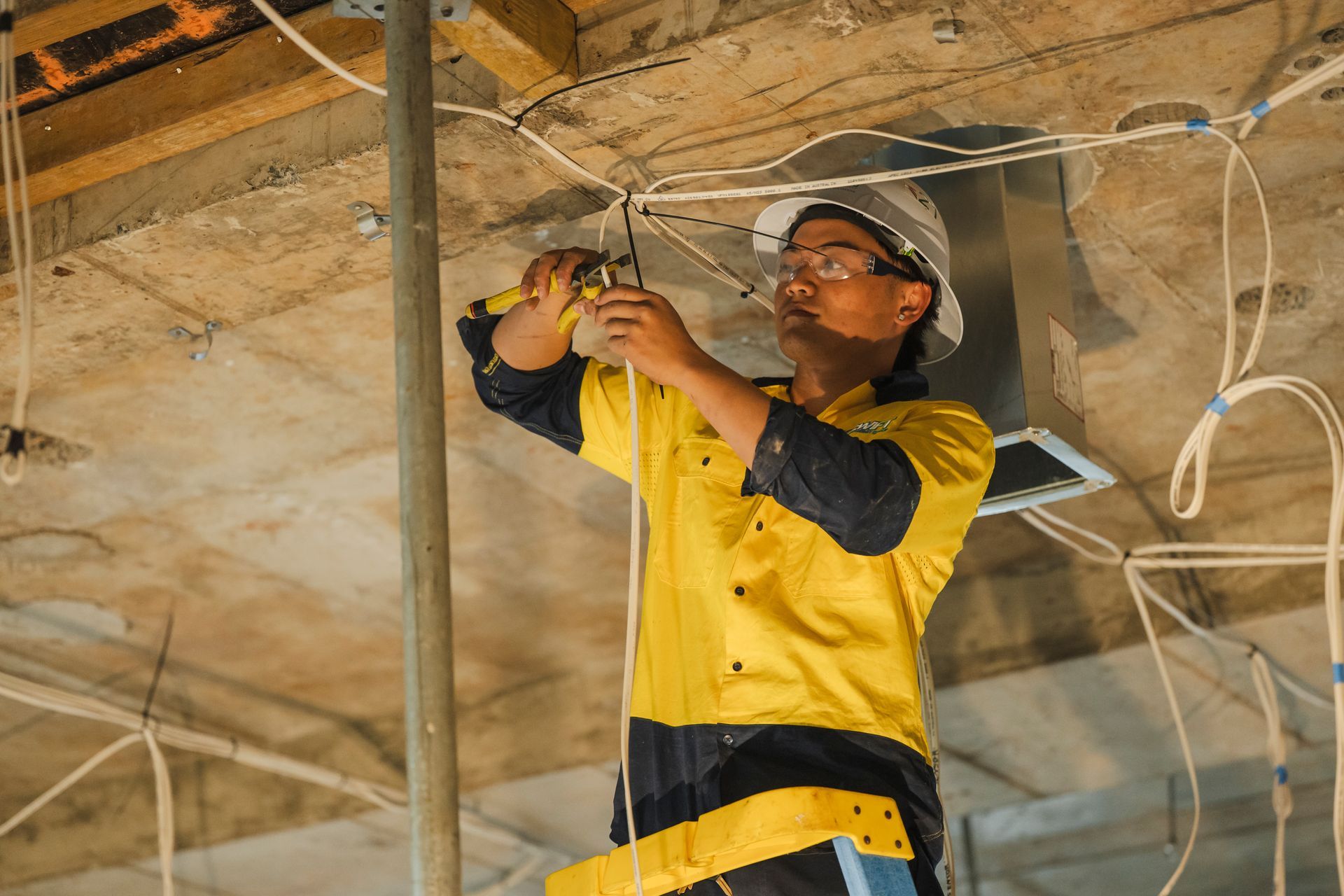 Electrician in yellow uniform, safety glasses, and hard hat, standing by orange machinery.