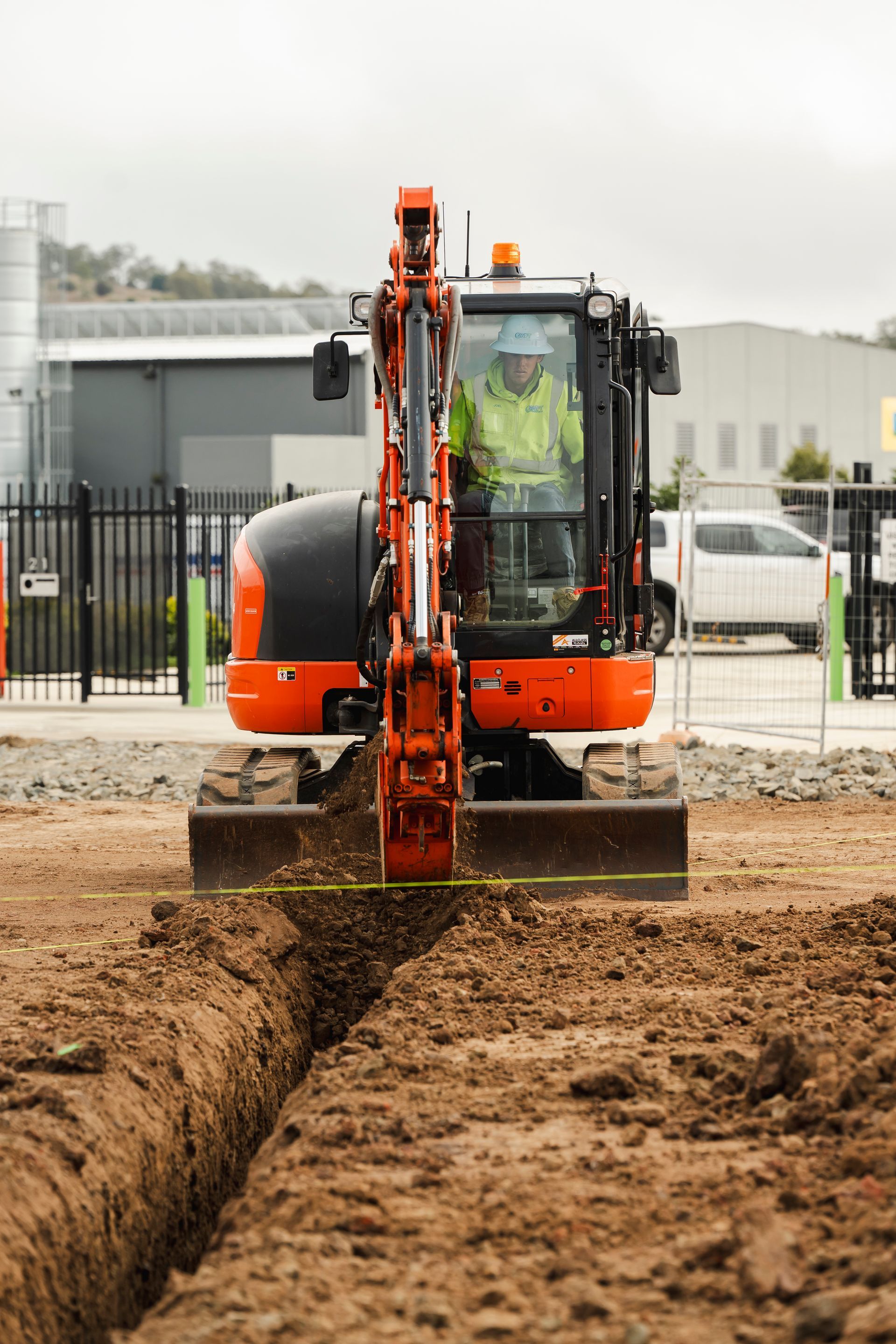 Orange excavator digging a trench, operator visible, in a construction site.