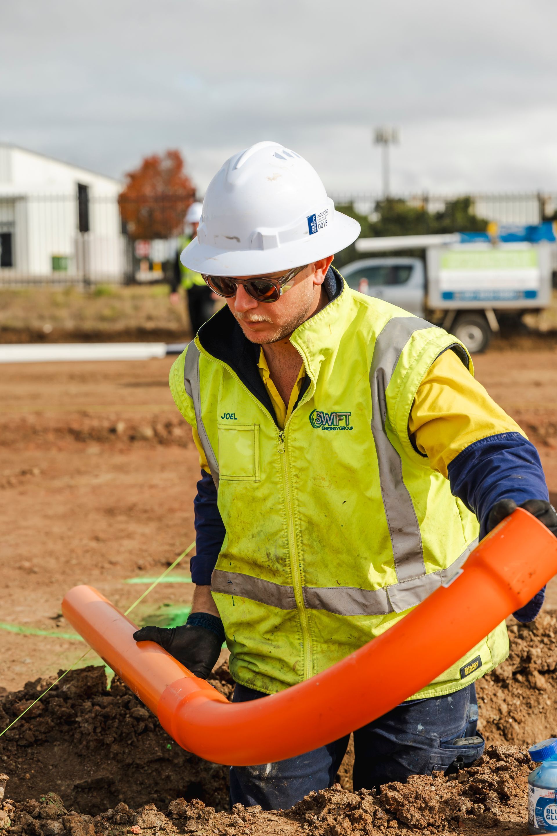 Electrician in hard hat and vest installs orange pipe in a dirt trench.