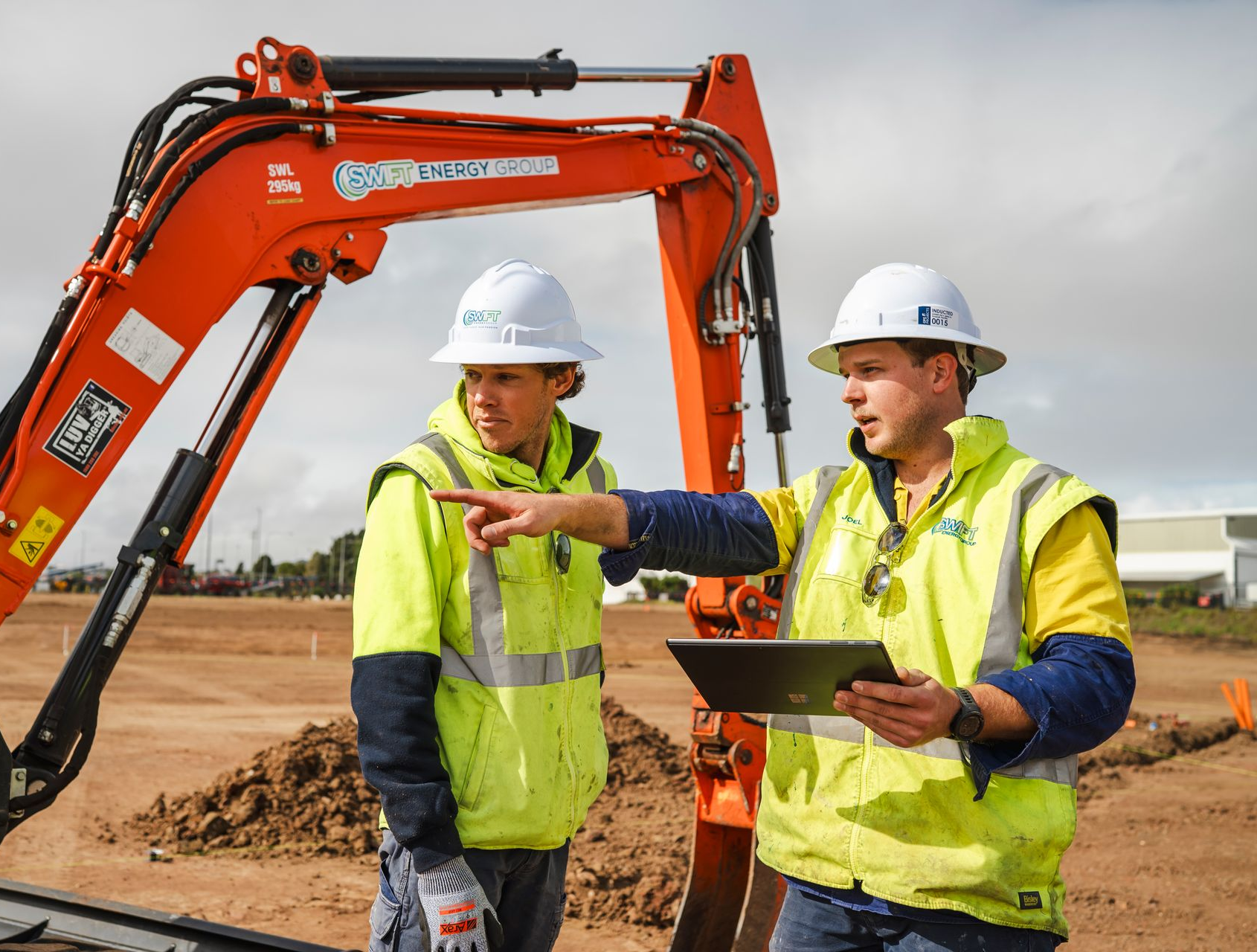 Two construction workers reviewing a tablet next to an orange excavator on a construction site.