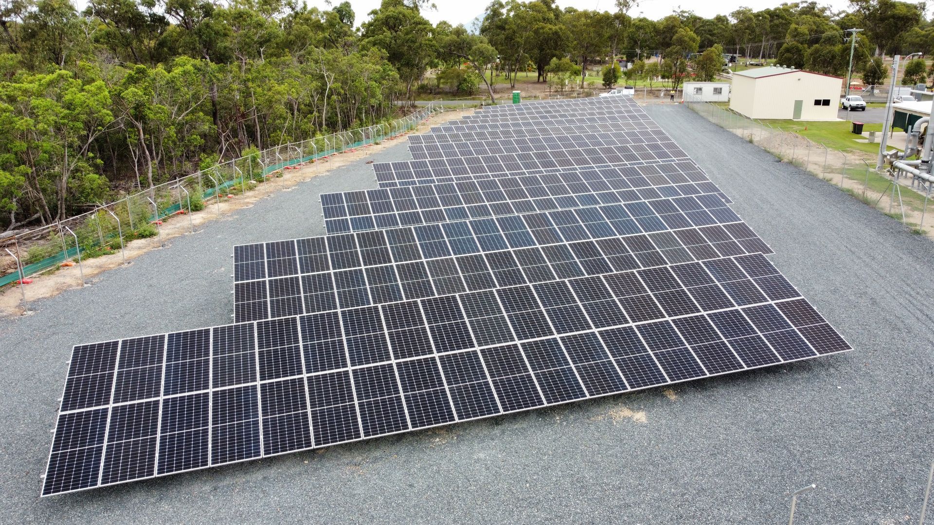 Solar panels on a gravel lot, arranged in rows, with trees and a building in the background.