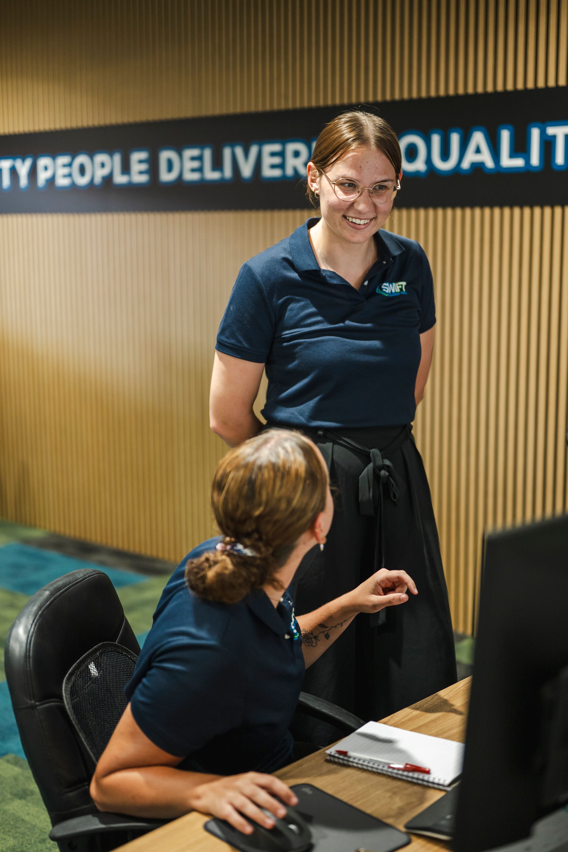 Two coworkers in matching navy uniforms confer at a computer station in an office with wood-paneled walls.
