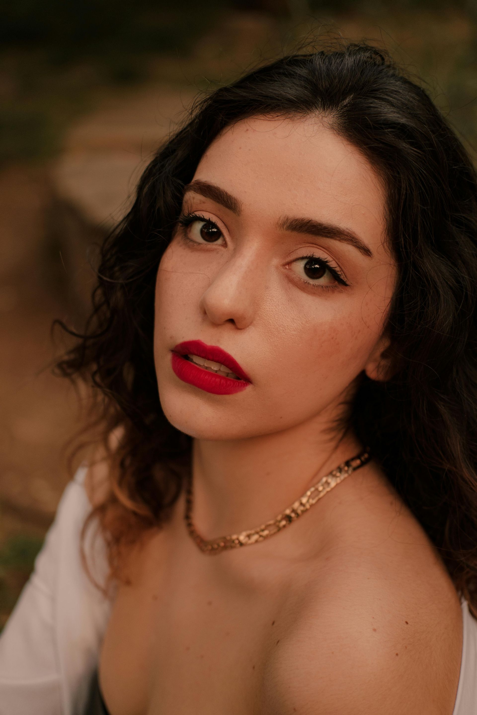 Woman with red lipstick and gold necklace looking at the camera outdoors.