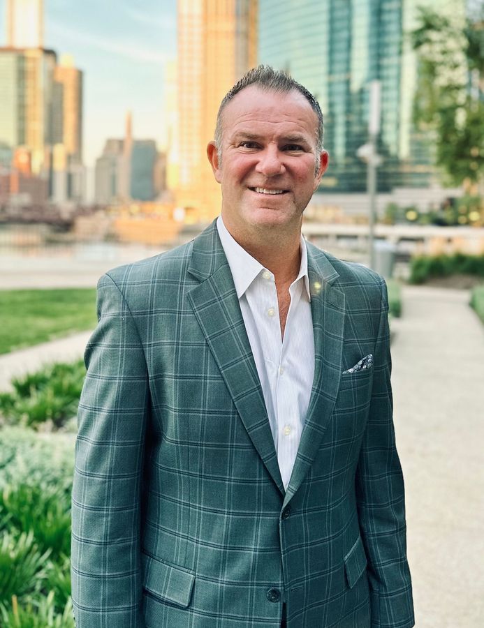 Man in a gray plaid blazer, smiling. Outdoors in a city setting with skyscrapers in the background.