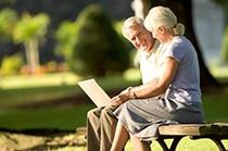 Elderly couple smiling at laptop on park bench, sunny day.