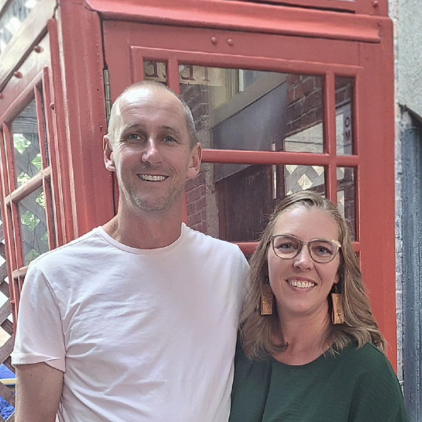 A man and a woman are posing for a picture in front of a red phone booth.