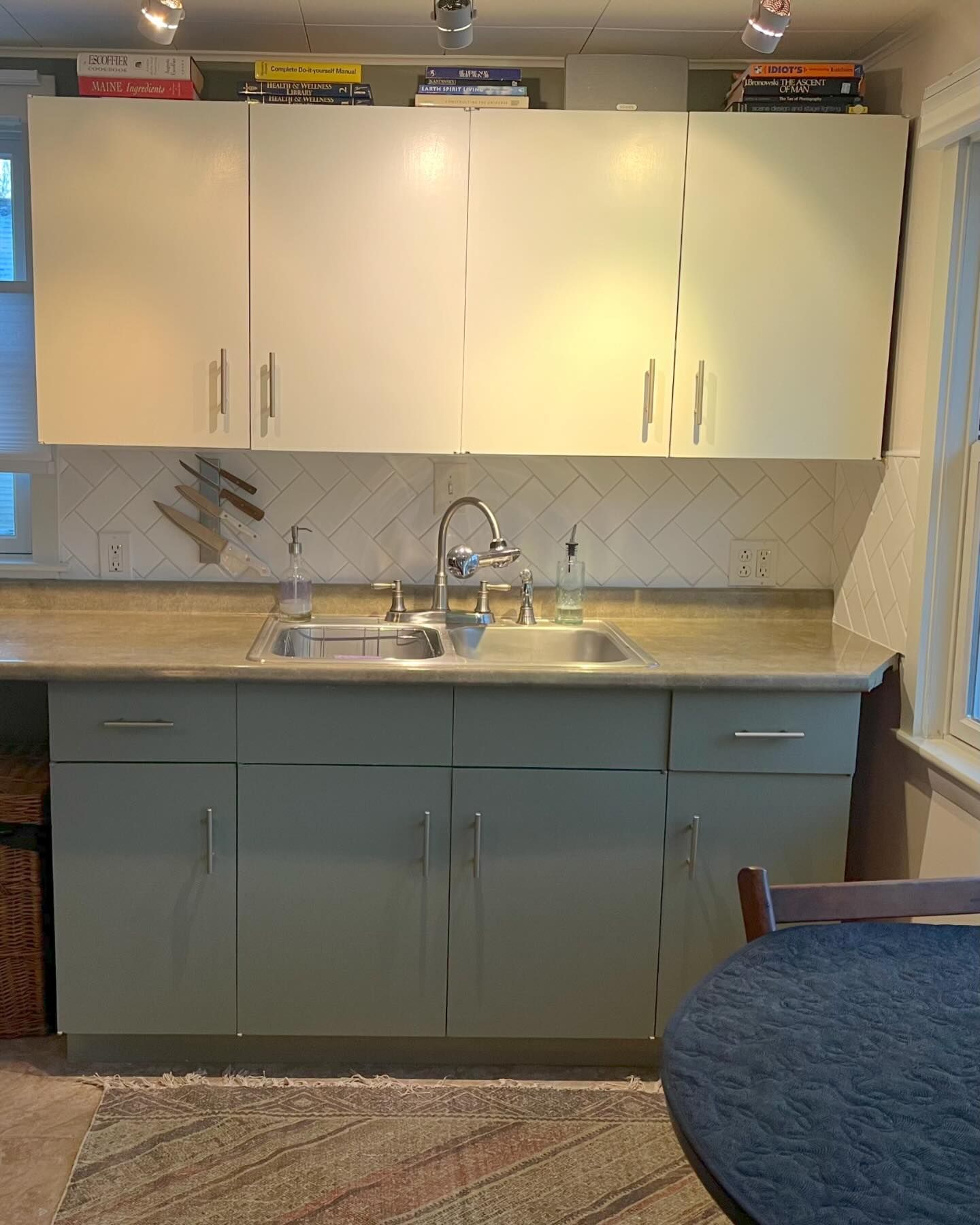 Kitchen with sage green cabinets, light countertops, and double sink. Books on upper white cabinets.