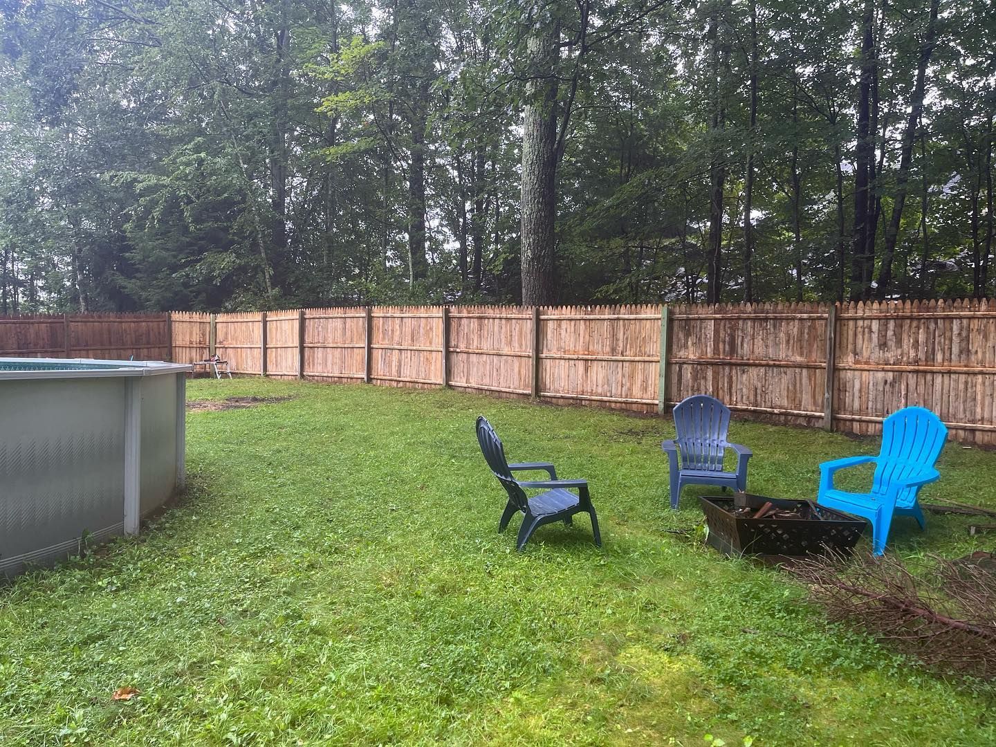 Backyard with a pool and fire pit, three chairs. A wood fence and trees are in the background. Overcast day.