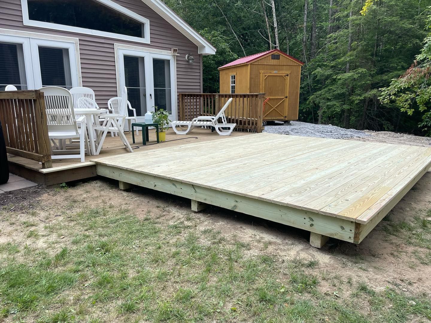 A wooden deck outside a house, with white chairs, a small shed, and grassy yard.