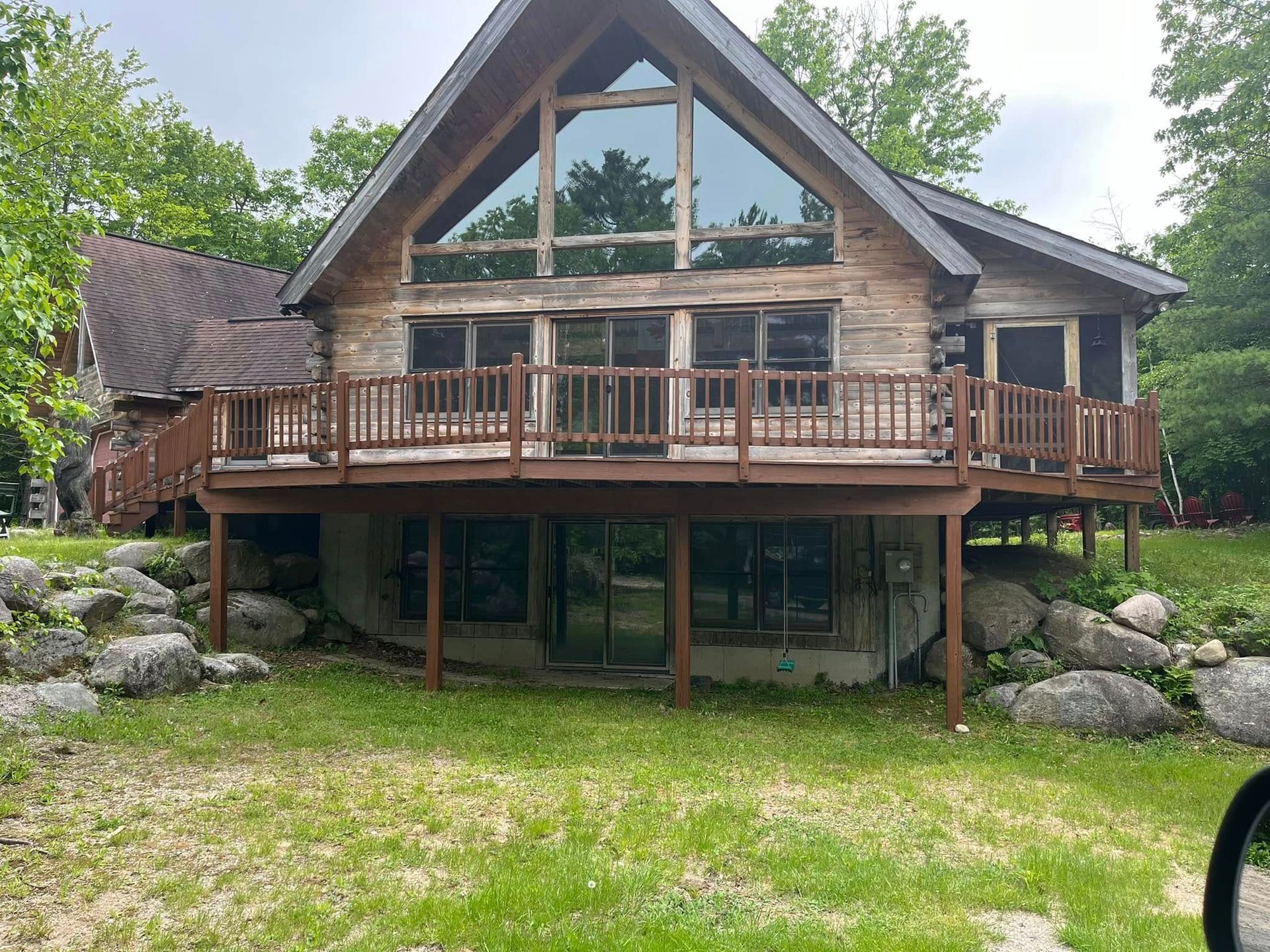 Log cabin with a large window and wraparound deck, set on a grassy area with exposed rocks.