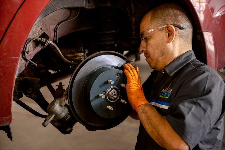Mechanic inspecting a car brake rotor inside a wheel well, wearing orange gloves and glasses