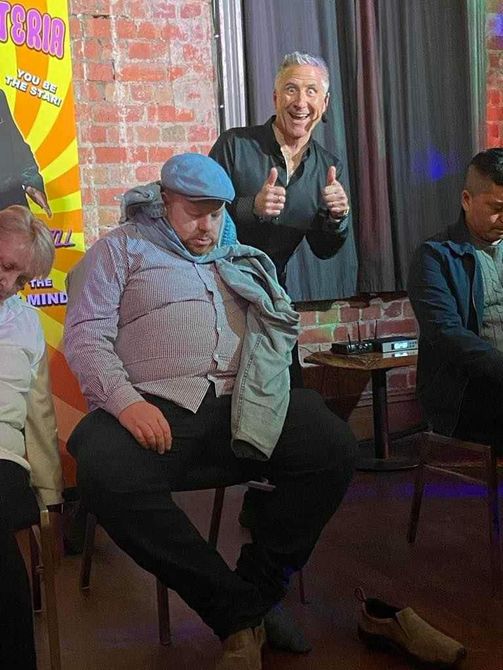 Man in blue cap and vest sits, another man gives thumbs up behind him at a venue.