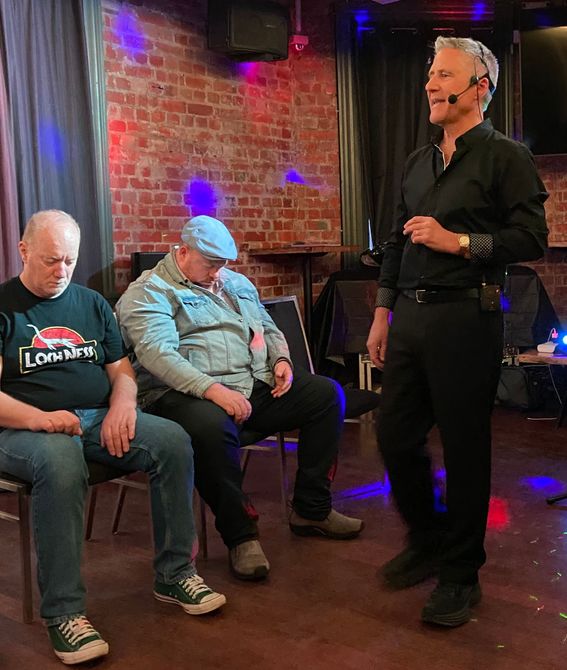 A man hypnotizes two seated men in a dimly lit room with a brick wall.