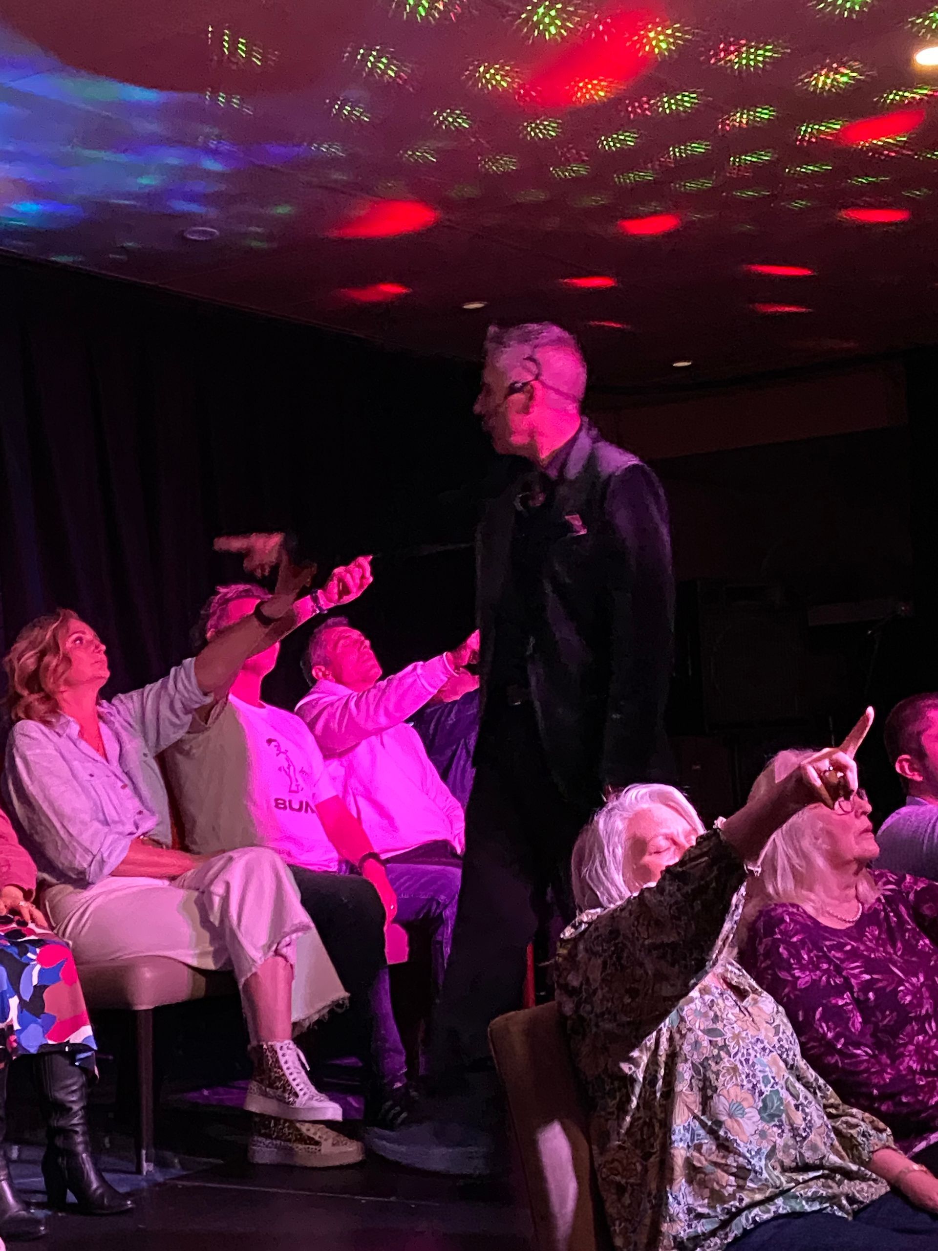 Man onstage with arms raised, people in audience point up, under red and green lights.