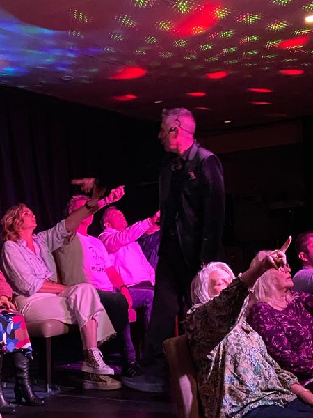 Man onstage with arms raised, people in audience point up, under red and green lights.