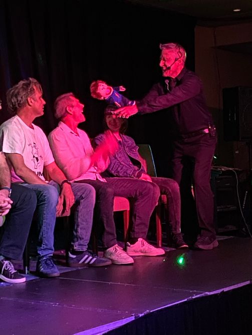Magician with puppet entertaining a seated audience on a stage.