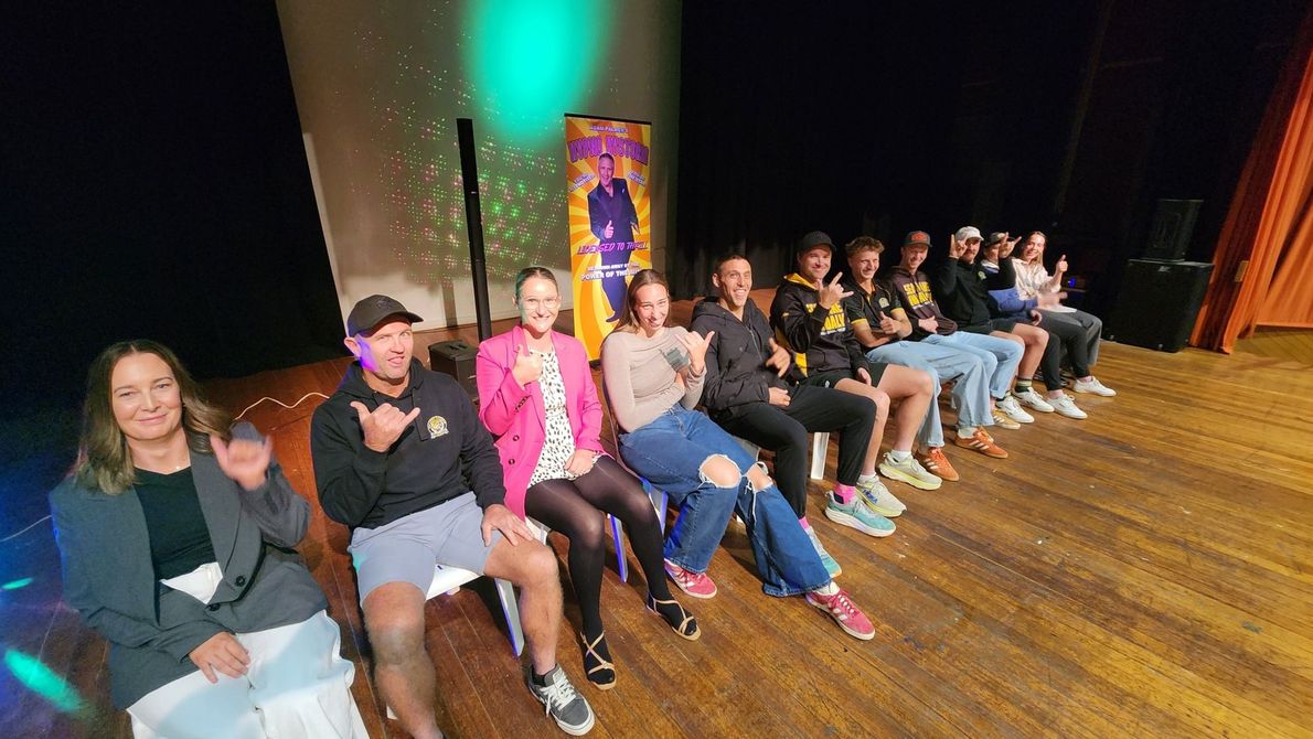 Group of people sitting onstage, some waving, with a banner behind them.